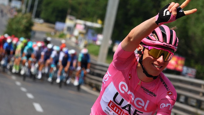 Tadej Pogacar porte le maillot rose du meneur et fait un signe victorieux de la main en pédalant, sourire aux lèvres.