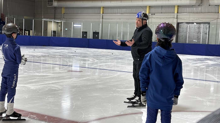 Homme debout sur la glace, parlant à deux jeunes patineurs.