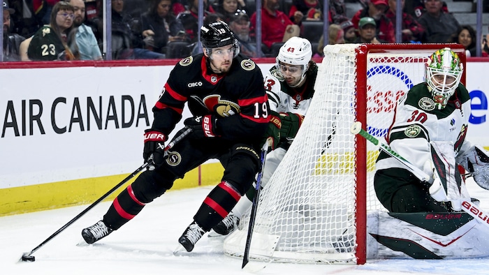 Un hockeyeur contourne le filet avec un adversaire à ses trousses pendant qu'un gardien s'agenouille.