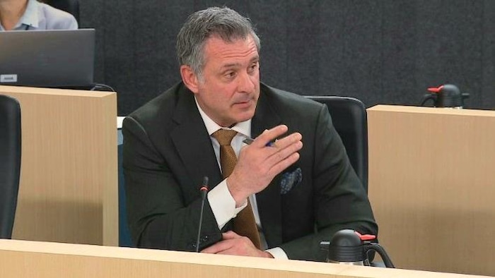 Un homme assis derrière un micro regarde à sa gauche d'un air sérieux et lève la main droite qui tient un stylo.