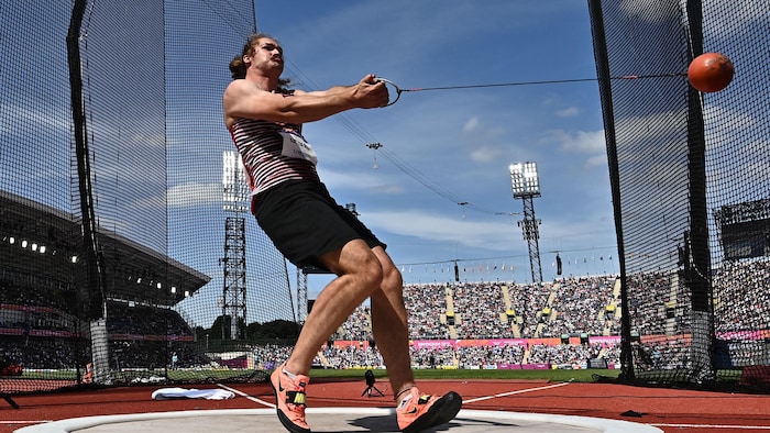 L'athlète, de profil, tient fermement à l'horizontal une boule accrochée à un fil de fer lors d'une rotation. 