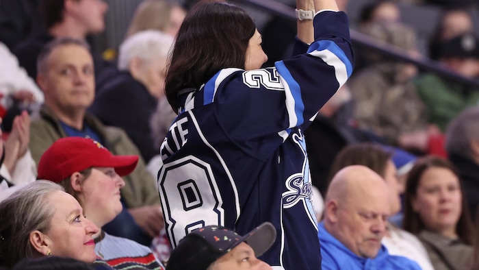 Dame debout avec un gilet des Saguenéens et qui applaudit. Les gens autour d'elle sont déçus.