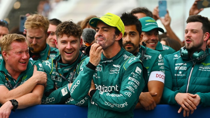 An F1 driver scratches his chin in front of his team's mechanics who laugh.  