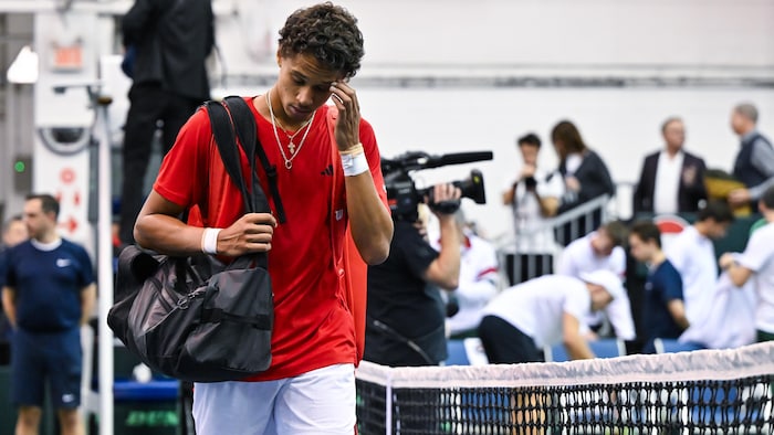 Un joueur de tennis marche le agelong d'un filet, en tenant un sac et avec une main sur le visage.