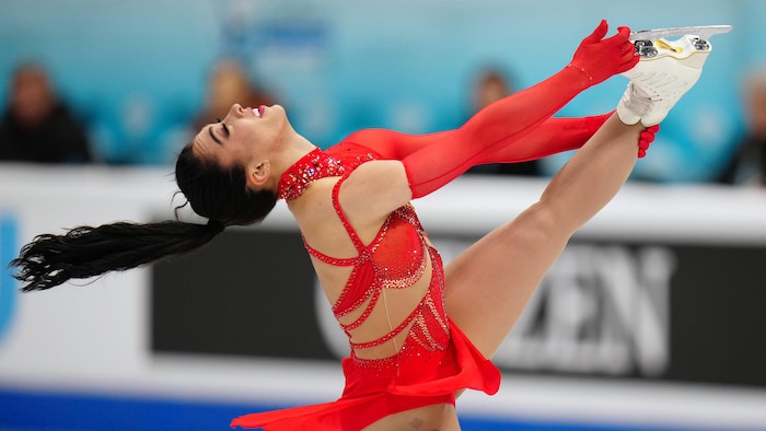 Une patineuse effectue une figure complexe où elle tient sa jambe par la cheville et la pointe du patin.
