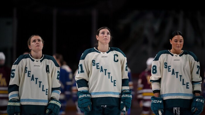 Trois joueuses de hockey en uniforme attendent le début de la rencontre sur la glace.