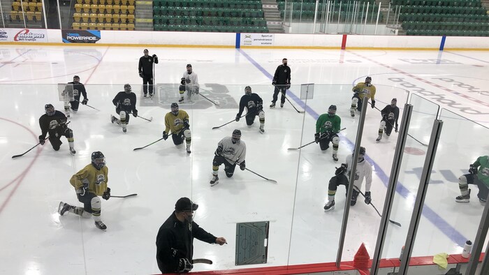 Des joueurs de hockey regarde leur entraîneur qui explique un exercice sur un tableau blanc. Ils portent tous un masque avec couvre-visage.