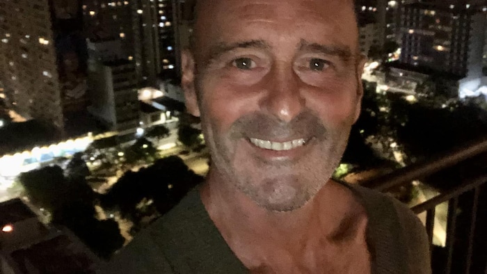 Un homme chauve, souriant, devant une ville, la nuit.