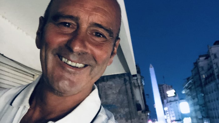 Homme chauve souriant. Derrière lui, la ville est illuminée, avec une rue grouillante d'activité.
