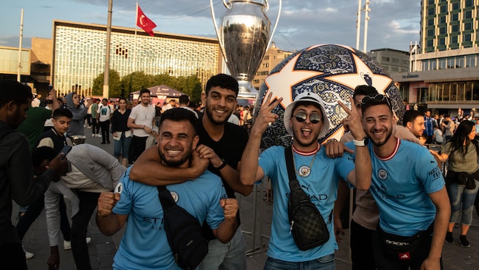 Des partisans avec des chandails aux couleurs de Manchester City posent pour la caméra devant un trophée et un ballon de soccer géants.