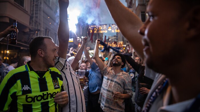 Des partisans de soccer festoient dans une petite rue avec un verre de bière à la main.