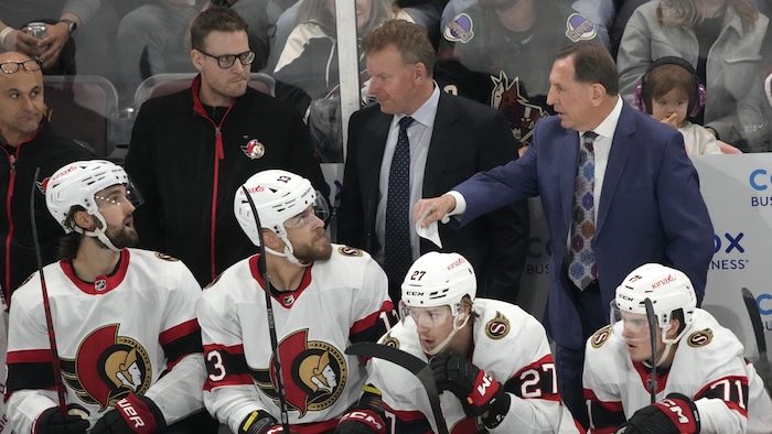 Derrière le banc de son équipe, un entraîneur de hockey tend le bras droit et parle à ses joueurs qui l'écoutent.