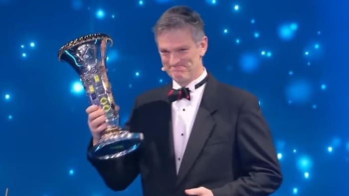 Un homme en habit de soirée sourit avec un trophée dans la main droite.