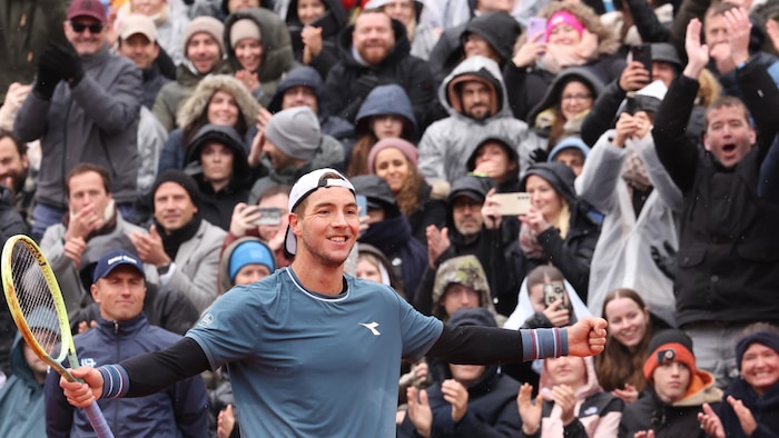 Un joueur de tennis sourit et tend les bras, avec sa raquette dans la main droite. La foule l'applaudit.   