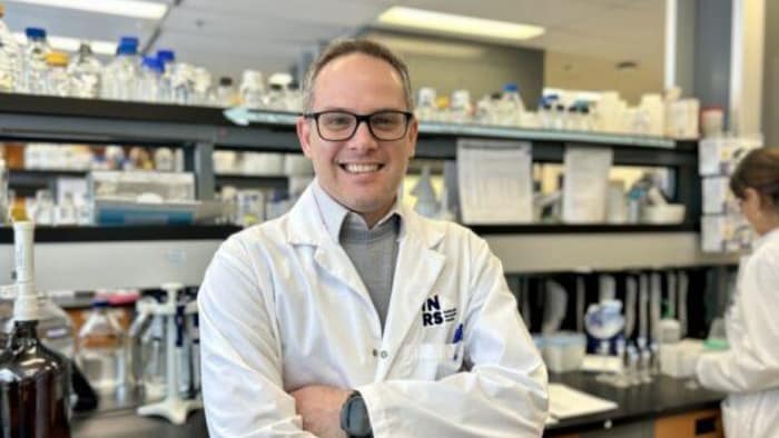 Un homme, avec une blouse blanche de scientifique, sourit dans un laboratoire.