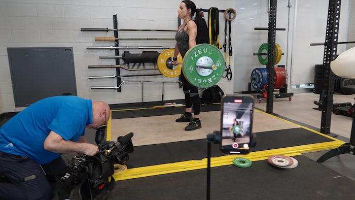 Josée Gallant tenant une barre avec des poids. Un iPhone est sur la droite tandis que le caméraman avec une autre caméra est accroupi devant elle.