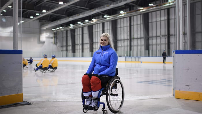 Une femme assise sur un fauteuil roulant sourit alors qu'un entraînement de parahockey se déroule derrière elle.