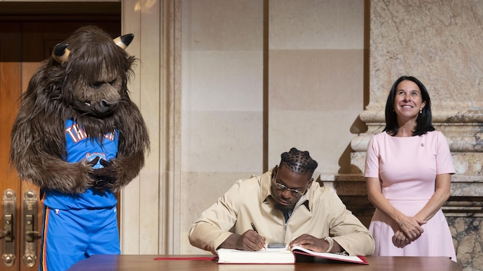 Un homme signe un livre d'or entre une mascotte et une femme qui regarde dans les airs. 