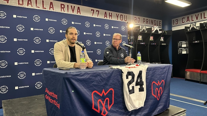 Les deux hommes sont assis derrière une table sur laquelle reposent deux micros et un chandail de football numéro 24.