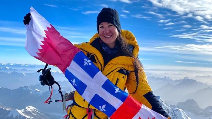 Une alpiniste au sommet d'une montage tient dans ses mains les drapeaux du Qatar, du Québec et du Canada