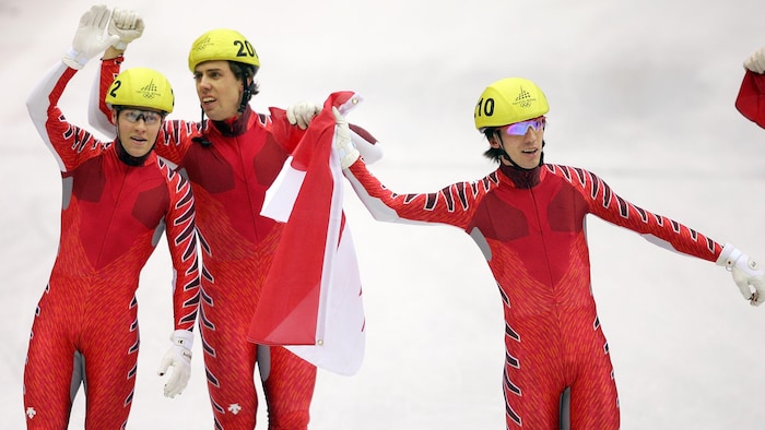 Un patineur de vitesse tient le drapeau canadien, tandis que ses deux coéquipiers saluent la foule.