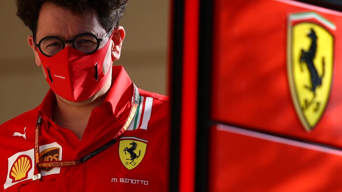 Un homme avec des lunettes et un masque sanitaire, en uniforme, marche droit devant lui, avec à sa gauche un panneau portant l'emblème de Ferrari.