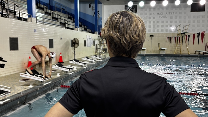 Jeune homme accroupi, prêt à se jeter à l'eau sous le regard de son entraîneuse, qui est de dos à la caméra.