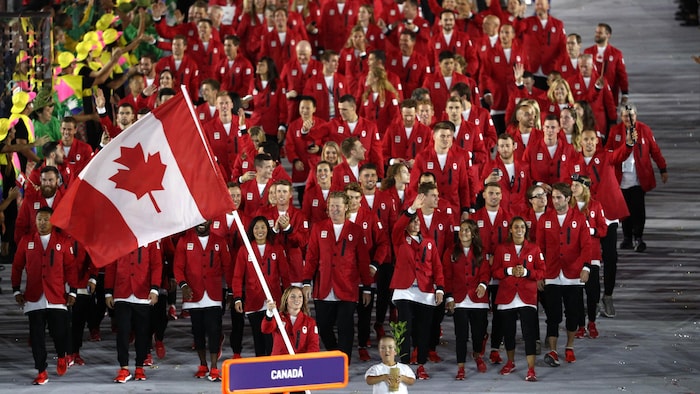Des hommes et des femmes habillés aux couleurs du drapeau canadien marchent en groupe, sourient et saluent la foule.