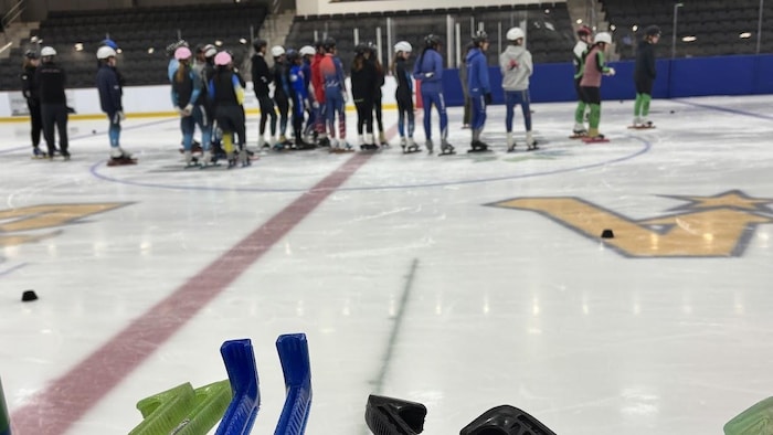 Jeunes patineurs debout, sur la glace, au loin. En avant-plan, des protège-lames sont sur le rebord de la bande. Une feuille avec des notes traîne avec les protège-lames.