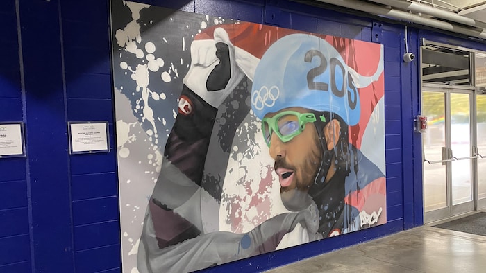 Sur un tableau, un patineur de vitesse lève un drapeau canadien au-dessus de son casque.