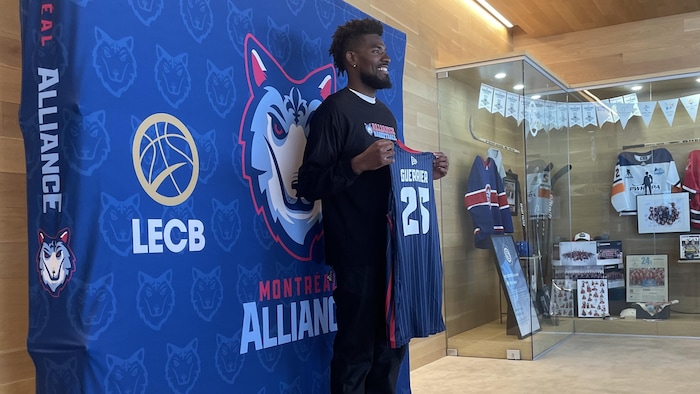 Il tient fièrement un chandail de l'Alliance en souriant.