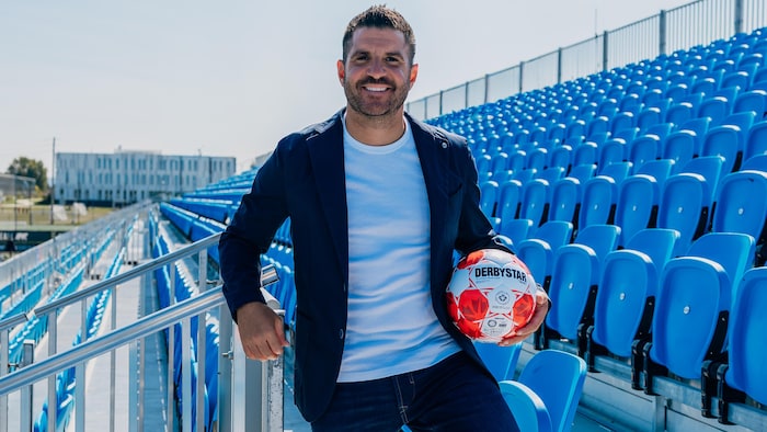 A man in empty stands holds a soccer ball in his left hand.