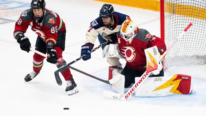 Deux hockeyeuses veulent la possession de la rondelle près d'une gardienne.