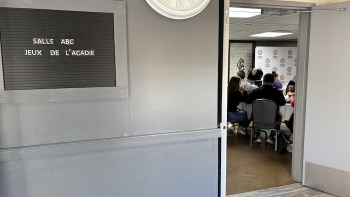 Écriteau avec Salle ABC Jeux de l'Acadie, à côté d'une porte ouverte où on entrevoit des gens assis.