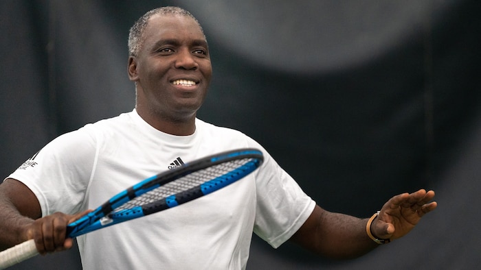 Un joueur de tennis grisonnant sourit et écarte les bras avec sa raquette dans la main droite.