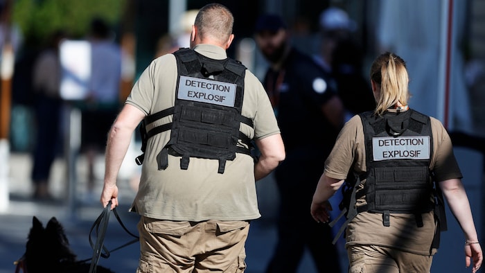 Un homme et une femme, vus de dos. Ils portent une veste où il est inscrit : détection explosif. L'homme tient un chien renifleur en laisse.