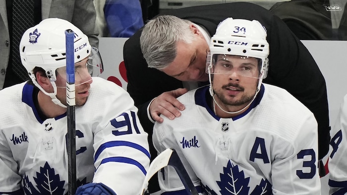 Un entraîneur de hockey parle à l'oreille d'un de ses joueurs assis au banc. Un coéquipier les regarde.