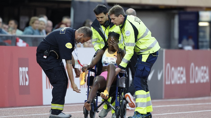 Une femme, entourée d'une équipe médicale, se tient la tête, assise sur un fauteuil roulant, le pied gauhce surélevé.