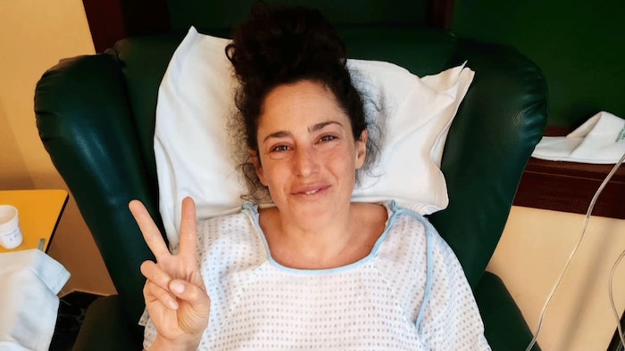 Elle fait un signe de la main et sourit depuis un fauteuil d'hôpital.