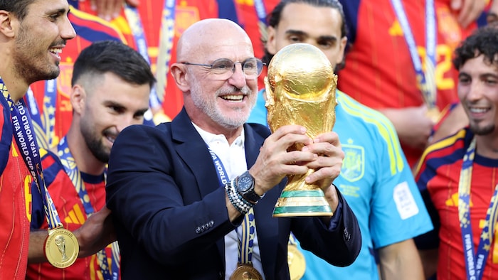 Il tient le trophée de la Coupe du monde devant ses joueurs.