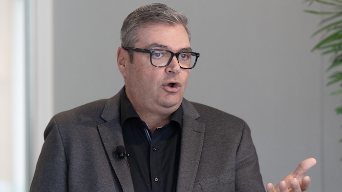 Il parle et fait un geste de la main gauche.