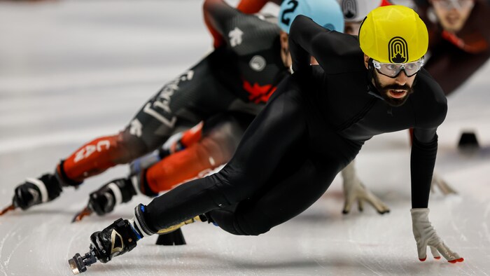 Un patineur de vitesse, vêtu d'une combinaison noire et d'un casque jaune, se penche vers la gauche pour effectuer un virage. 