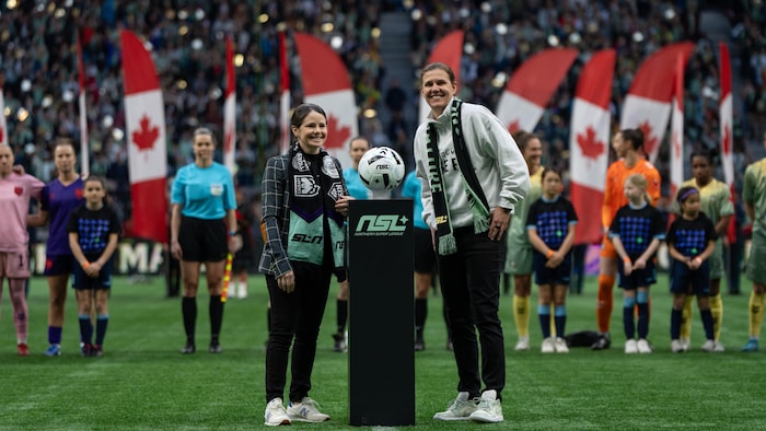 Devant joueuses, jeunes filles et drapeaux canadiens, les deux femmes présentent un ballon au centre du terrain.