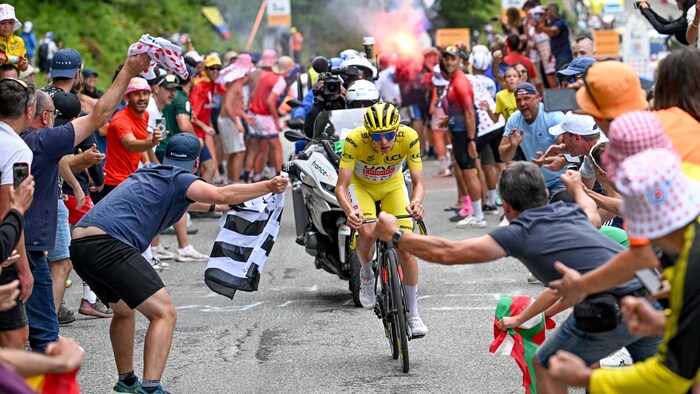 Un cycliste roule sur une route, il est entouré de spectateurs, une moto avec un caméraman se trouve derrière lui.