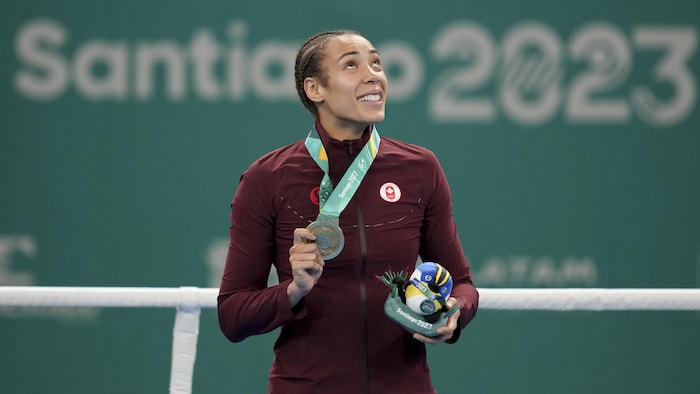 Boxer Tamara Thibault holds the medal around her neck in her hand as she looks skyward on the Pan American Games podium in the ring in front of the Santiago 2023 sign.