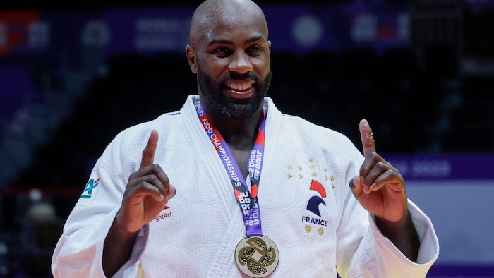 Un judoka souriant lève ses deux index en signe de victoire, avec une médaille d'or au cou.