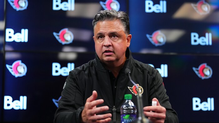 Un entraîneur de hockey répond aux questions des journalistes pendant une conférence de presse, devant un écran avec le logo de son équipe et de son commanditaire.