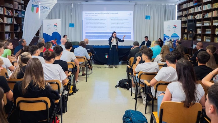 Elle tient un micro et parle à des étudiants assis devant elle.
