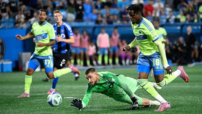 Un gardien de but au shot tente d'attraper le ballon alors qu'il est entouré de deux adversaires.