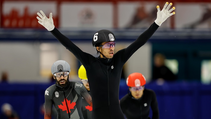 Un patineur de vitesse, vêtu d'une combinaison toute noire, célèbre sa victoire en levant les bras après une course. 